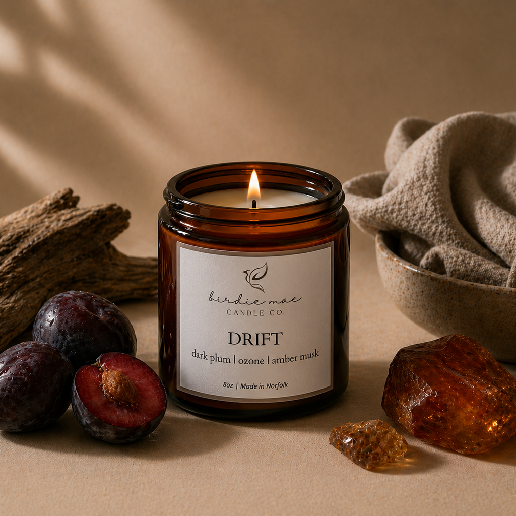 A lit amber glass jar candle labeled 'birdie mae CANDLE CO DRIFT' sits on a beige surface with dark plums, driftwood, a folded linen cloth, and amber stones around it.