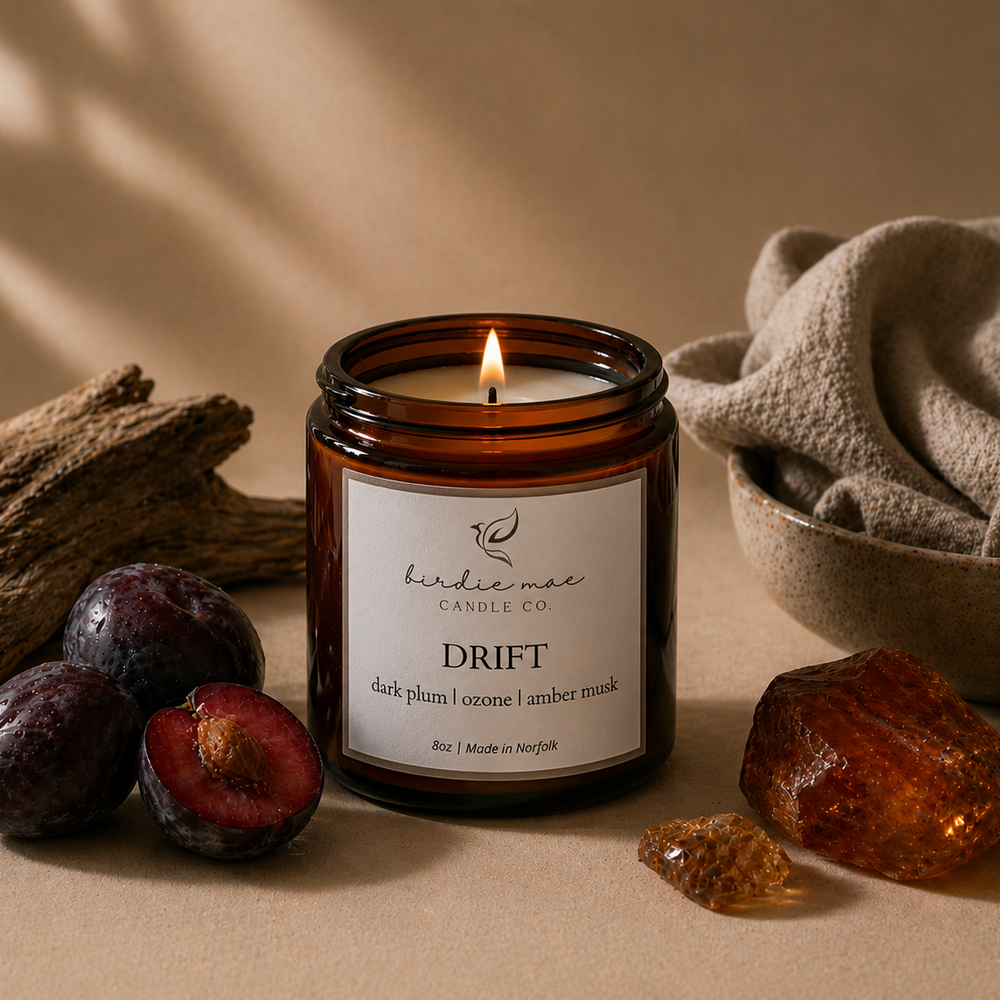 A lit amber glass jar candle labeled 'birdie mae CANDLE CO DRIFT' sits on a beige surface with dark plums, driftwood, a folded linen cloth, and amber stones around it.
